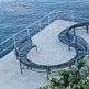 Curved outdoor metal benches arranged on a concrete platform overlooking a blue ocean, with white railings and some greenery in the foreground.