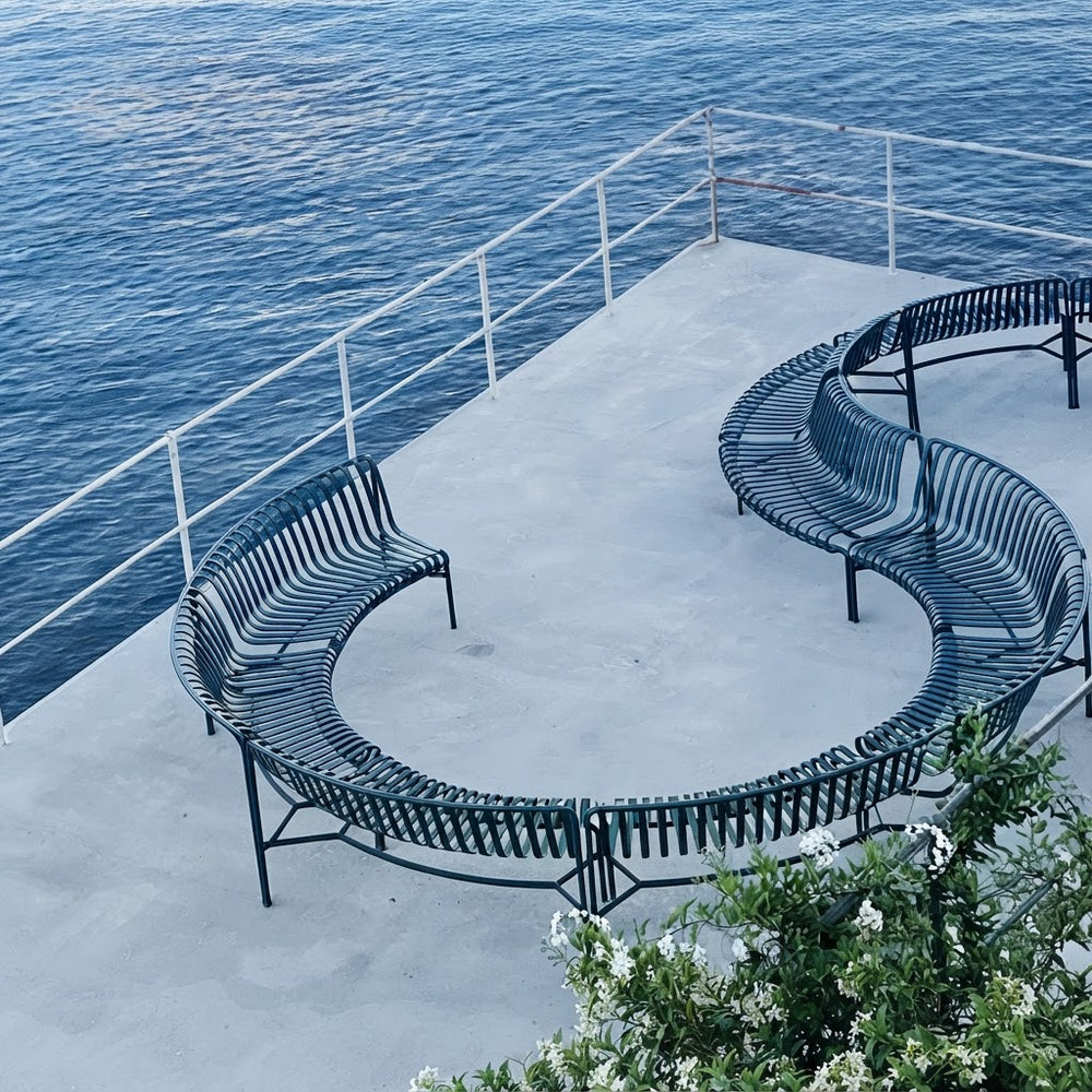 Curved outdoor metal benches arranged on a concrete platform overlooking a blue ocean, with white railings and some greenery in the foreground.