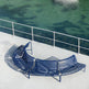 A curved outdoor metal bench in dark blue sits on a light concrete deck beside a railing overlooking greenish water.