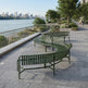 Three curved green outdoor benches arranged on a paved waterfront promenade with a river, trees, and city skyline in the background.
