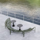 Outdoor curved metal patio seating set with two small round tables, placed on a concrete deck beside a body of water with a white railing.