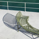An olive green metal lounge chair with a curved, slatted design, positioned on a white deck with a white railing and dark teal water in the background. The chair is cast a distinct shadow to the left.