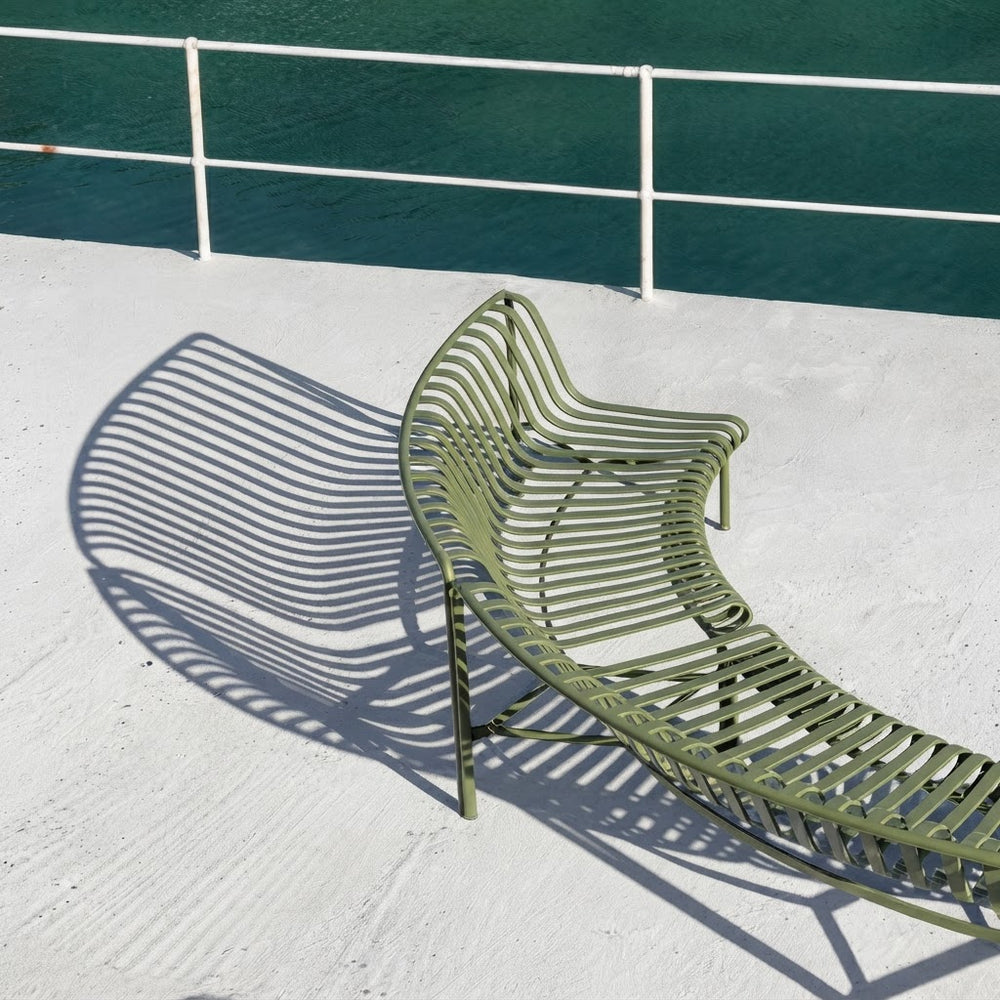 An olive green metal lounge chair with a curved, slatted design, positioned on a white deck with a white railing and dark teal water in the background. The chair is cast a distinct shadow to the left.
