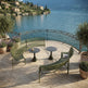 Outdoor curved metal patio seating set with two olive green lounge chairs and two small round tables on a stone terrace overlooking a scenic lake with buildings and hills in the background.
