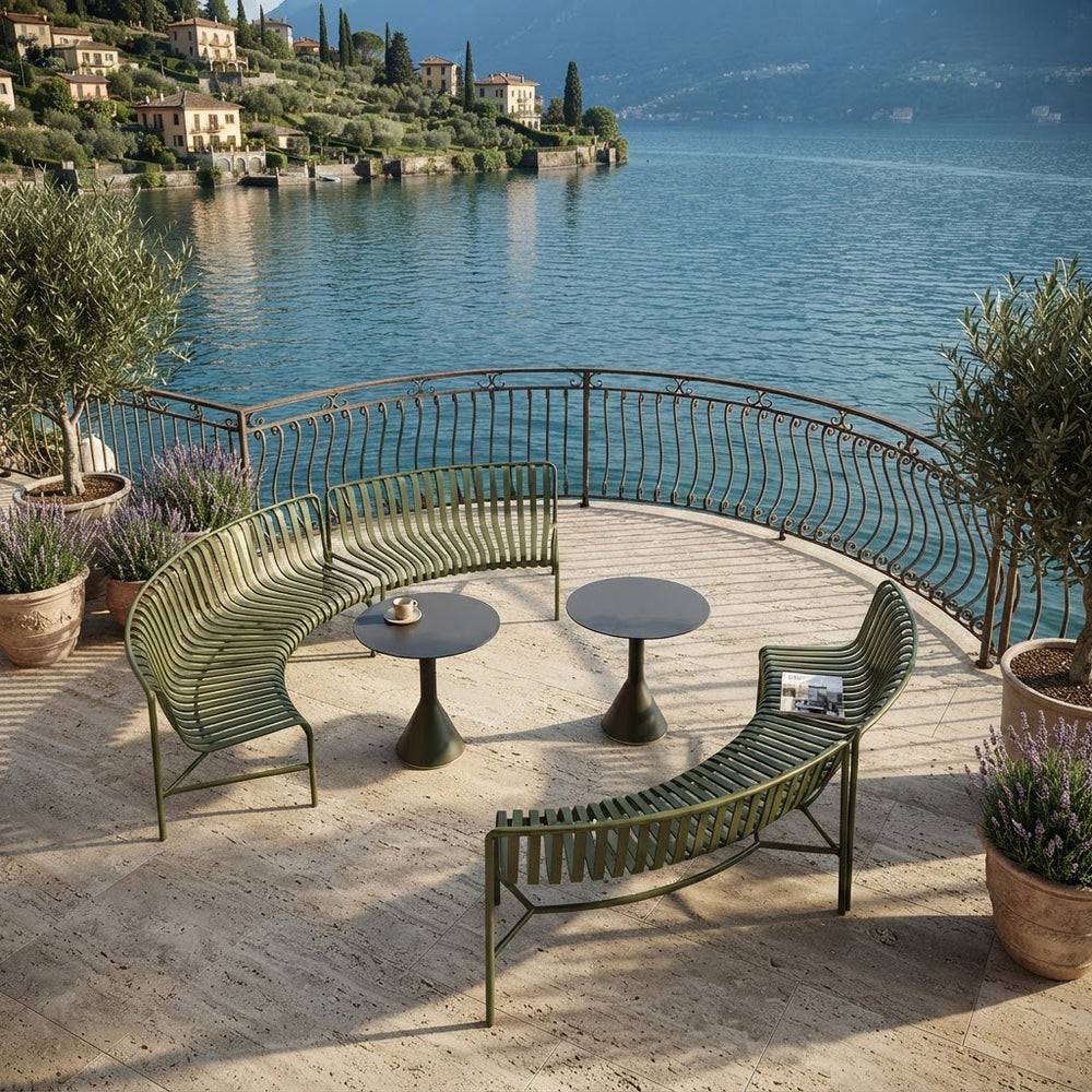 Outdoor curved metal patio seating set with two olive green lounge chairs and two small round tables on a stone terrace overlooking a scenic lake with buildings and hills in the background.