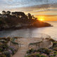 A scenic coastal sunset view with a curved stone patio, metal railing, and two green benches facing the ocean. Rocky shoreline, trees, and flowers frame the scene under a glowing sun and dramatic clouds.
