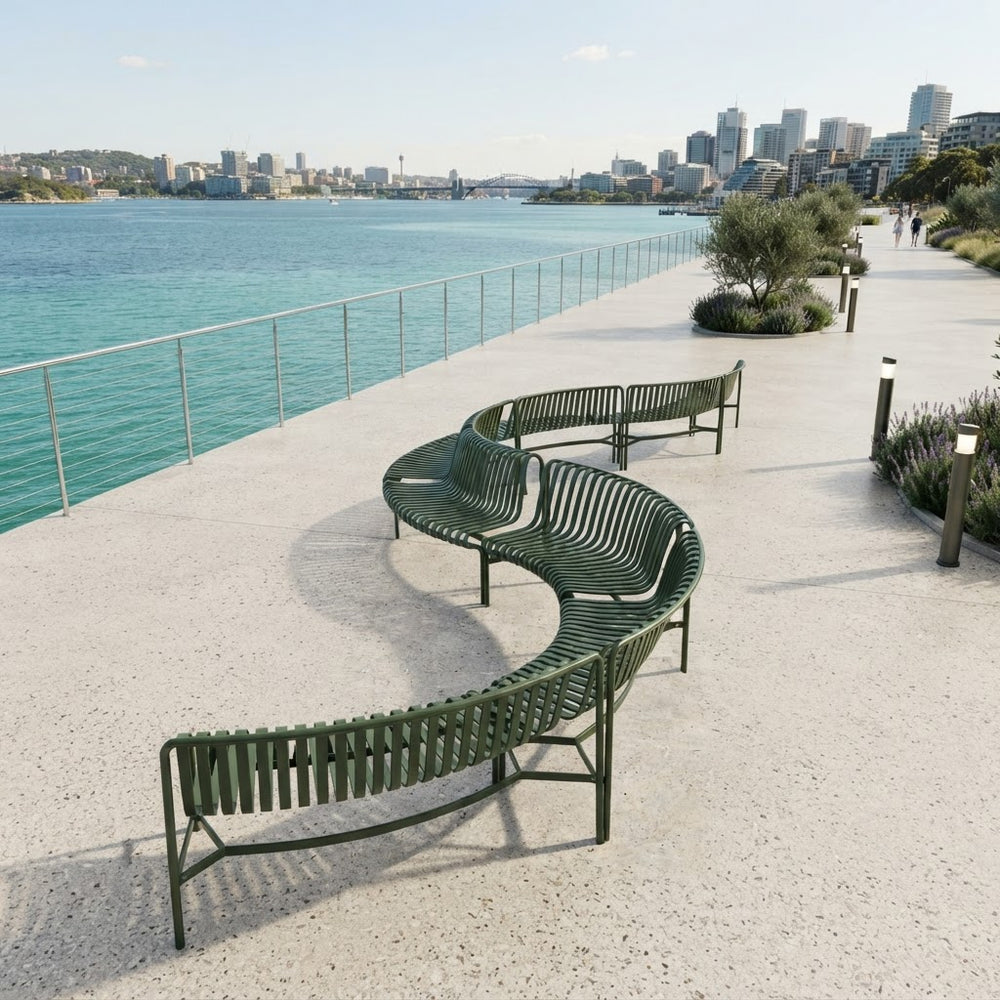 A curved outdoor metal bench in dark green sits on a light concrete waterfront promenade with a railing, blue water, and a city skyline in the background.