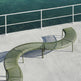 A curved outdoor bench with olive green metal slats sits on a light concrete deck beside a railing overlooking blue-green water.
