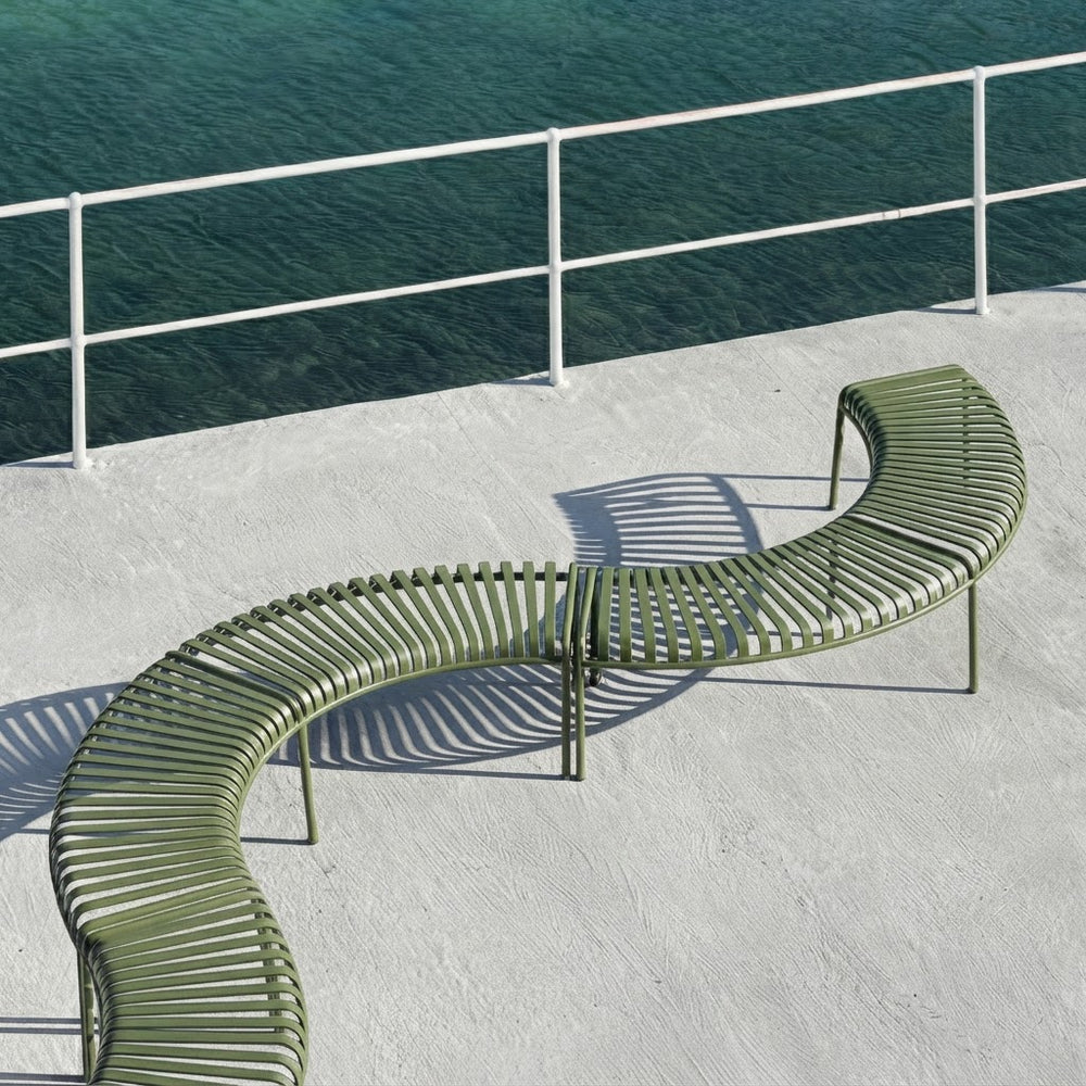 A curved outdoor bench with olive green metal slats sits on a light concrete deck beside a railing overlooking blue-green water.