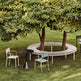 Outdoor patio furniture arrangement on a grassy lawn beneath a large leafy tree, featuring a curved white slatted bench, a small black round table, and two chairs in off-white and pale mint green.