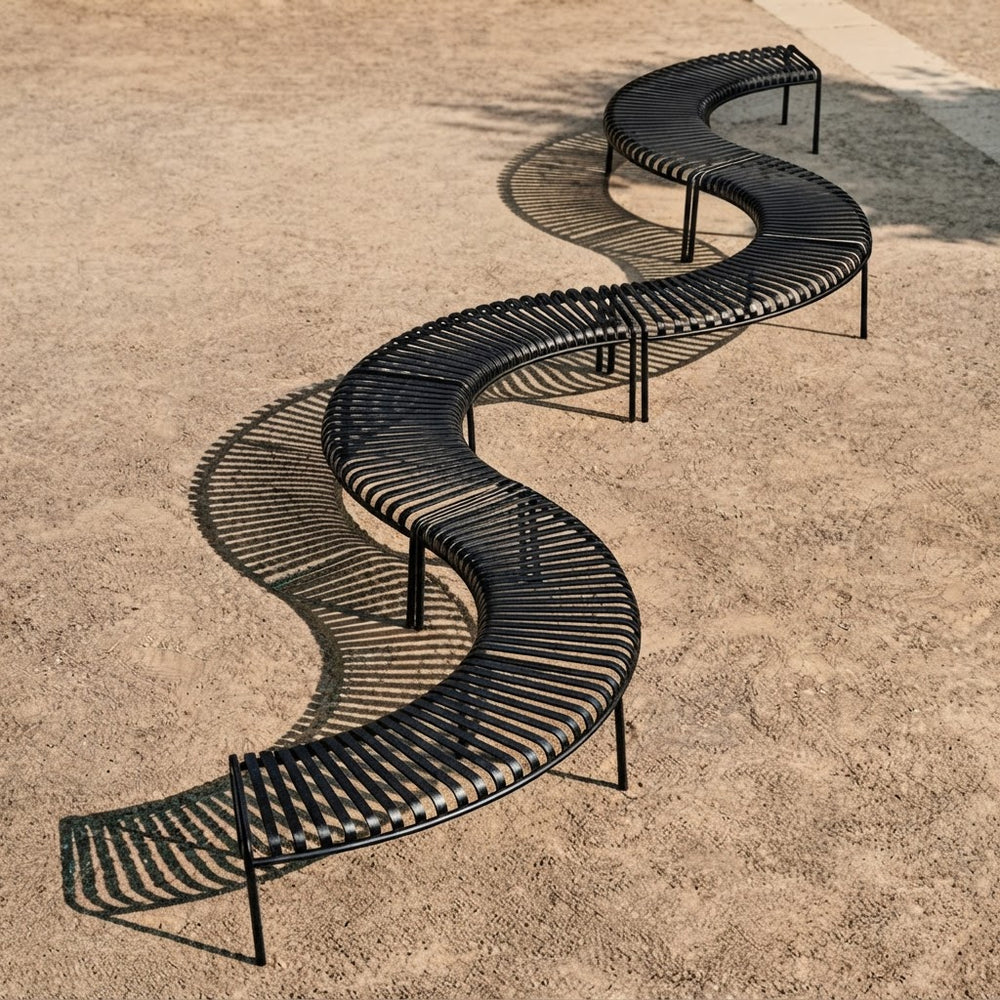 A long black curved metal bench with a slatted design, arranged in a serpentine S-shape on a sandy ground. The bench casts a prominent shadow on the light brown sand.