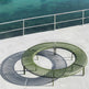 An outdoor circular metal lounge bench in olive green sits on a light concrete deck beside a railing overlooking clear turquoise water. The bench has a slatted curved design with visible support legs, and its shadow is cast sharply on the deck.