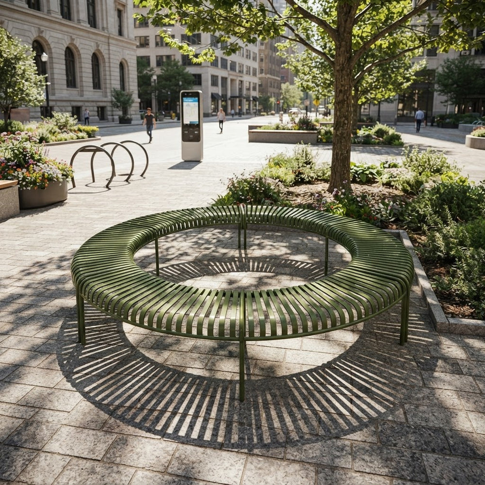 A circular outdoor bench made of green metal slats sits in a city plaza with stone paving, trees, planters, and buildings in the background.