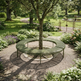 A round green slatted bench arranged around the base of a tree in a landscaped garden with gravel ground cover, plants, and a fountain visible in the background.