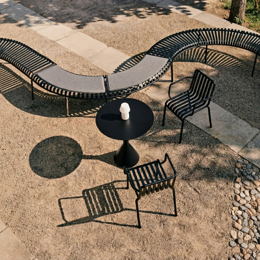 Modern outdoor patio furniture set featuring a curved black metal bench with gray cushions, two matching black metal armchairs, and a round black pedestal table on a stone and gravel patio.