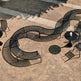 A curved black metal outdoor lounge bench with a wavy, serpentine design, shown from an elevated angle on a sandy patio with concrete walkways and casting a long shadow.