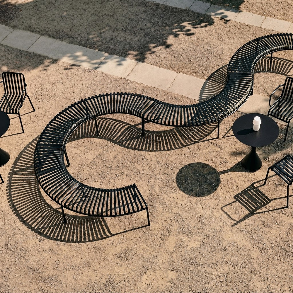 A curved black metal outdoor lounge bench with a wavy, serpentine design, shown from an elevated angle on a sandy patio with concrete walkways and casting a long shadow.