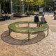 A large circular outdoor bench in a modern urban plaza, with several people sitting on it. The bench has a slatted olive green metal frame and is situated on a brick-paved circular area with gravel around it.
