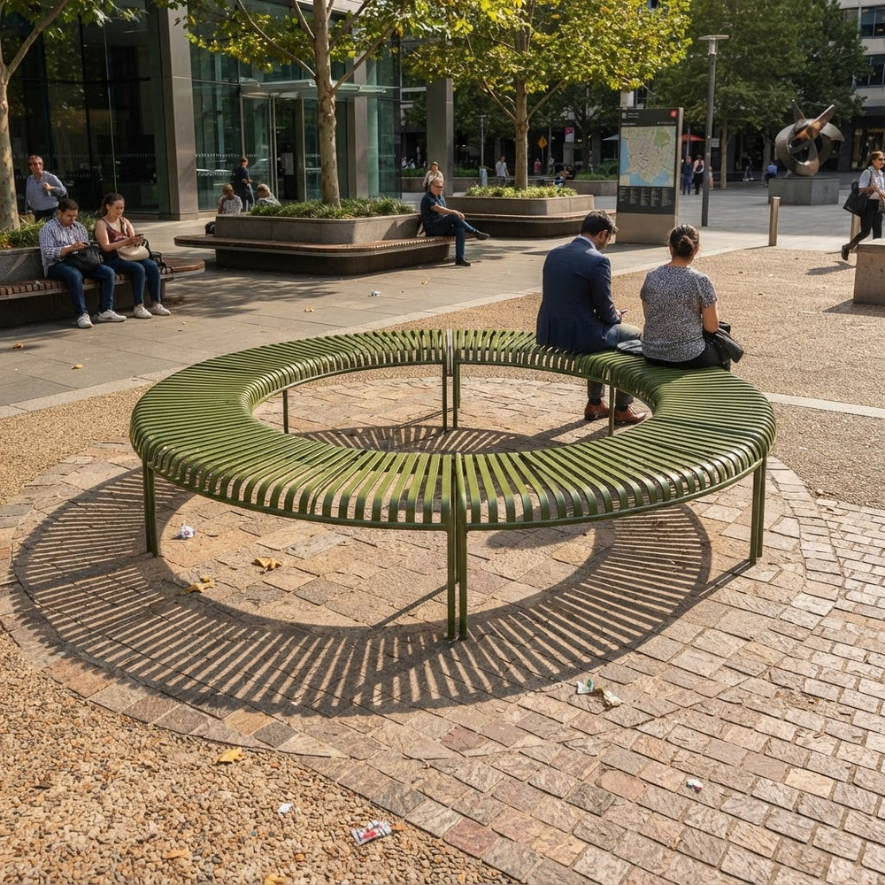 A large circular outdoor bench in a modern urban plaza, with several people sitting on it. The bench has a slatted olive green metal frame and is situated on a brick-paved circular area with gravel around it.