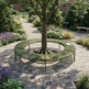 A circular outdoor bench made of green slats is arranged around the base of a tree in a landscaped garden courtyard with a stone paver floor, plants, and a walkway.
