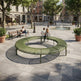 A large circular outdoor bench in olive green metal sits in a paved public square, with people sitting and walking around it. Trees, buildings, benches, and planters are visible in the urban setting.