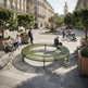 A large circular outdoor bench in olive green metal sits in a sunny urban plaza with people sitting and walking around it, surrounded by trees, buildings, and a fountain in the distance.