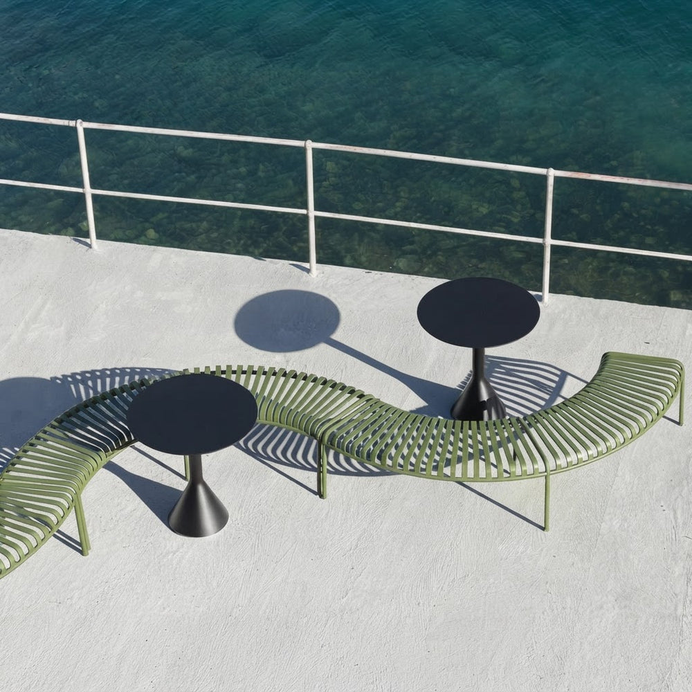 Outdoor curved lounge seating set with olive green slatted benches and round black tables on a white deck overlooking clear turquoise water.