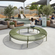 An outdoor circular slatted bench in muted olive green is shown in a modern playground seating area, with a child standing in the center and adults seated on nearby lounge chairs under a canopy.