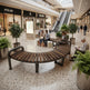 A curved wooden bench with dark metal armrests sits in a spacious, modern shopping mall atrium with terrazzo floors, potted plants, escalators, and shoppers sitting nearby.