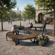 Curved wooden park benches with black metal frames in a paved urban plaza, with several people sitting and walking around them under trees.