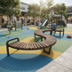 Outdoor playground scene with curved wooden benches, a slide, climbing wall, and children playing on colorful rubberized surfacing in front of retail buildings.
