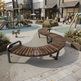 A curved outdoor wooden bench with dark metal armrests and legs, situated in a modern plaza with a children's playground, storefronts, and planters in the background.