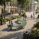 A curved green wooden bench in a paved urban plaza, with two people sitting on it and several pedestrians walking nearby. Trees, flower beds, buildings, and street lamps are visible in the background.