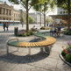 A curved outdoor bench with a green and light wood slatted seat and silver metal frame, placed in a paved public square with trees, flowers, and people sitting nearby.