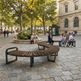 Curved wooden park benches with black metal frames in a cobblestone plaza, with several people sitting and walking nearby in a city park setting.