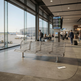 A modern three-seat metal bench with a slatted design sits in an airport terminal near large windows, with passengers walking and airplanes visible outside.