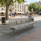 A modern stainless steel park bench with a curved slatted design, situated in a paved public square with people and historic buildings in the background.