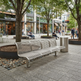 A modern three-seat stainless steel park bench in an outdoor urban plaza, shown with a matching cylindrical metal trash can and stone planters in a city setting with storefronts in the background.