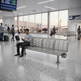 A man sits on a modern three-seat metallic airport waiting area bench, looking at his phone in a spacious, bright terminal with large windows, other travelers, and airplanes visible outside.