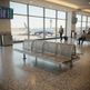 A modern metal airport waiting area with a curved bench in the foreground, large windows showing an airplane outside, and a few travelers seated nearby.