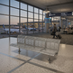 A modern three-seat stainless steel airport waiting room bench in the foreground, with a large glass window behind it showing airplanes and travelers at an airport terminal.