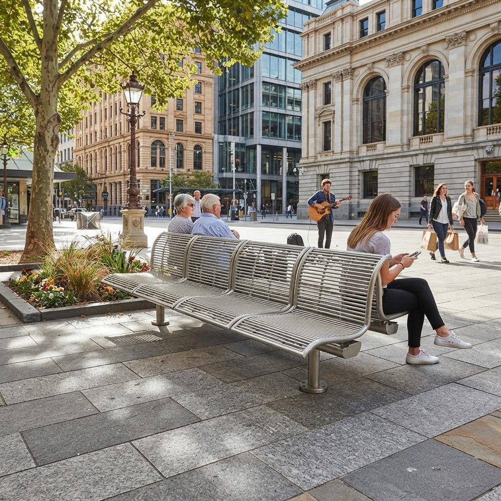 A modern curved metal bench in a city plaza, with people sitting and using it. The bench has a slatted design and is shown in an outdoor urban setting with buildings, trees, and pedestrians in the background.