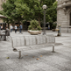 A modern three-seater metal bench with a slatted back and seat, situated in an outdoor urban plaza with people, trees, and stone buildings in the background.