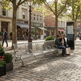 A modern curved metal bench in a cobblestone town square, with two people sitting on it and a map kiosk nearby. The scene is set in a real outdoor urban environment with trees, storefronts, and a lamppost.