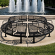 A circular black metal mesh bench seating area with multiple curved seats arranged in a ring on a concrete plaza, with a fountain and green lawn in the background.