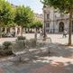 A modern three-seater metal bench with a curved, woven slat design sits on a paved plaza in a city square. The scene includes trees, historic buildings, and several pedestrians in the background.