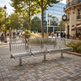 A modern three-seat stainless steel wire mesh bench in an outdoor urban plaza with paved stone flooring, trees, and pedestrians in the background.
