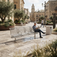 A man sits on a modern metal bench in a sunlit urban plaza, reading a newspaper. The bench has a woven slat design with integrated armrests and curved backrests.