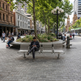 A man sits on a modern curved metal bench in a city plaza, looking at his phone. The scene includes other benches, people, trees, street lamps, and urban buildings in the background.