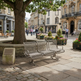 A modern three-seat metal bench with a woven wire backrest sits in a paved town square beside a large tree, with historic buildings and pedestrians in the background.